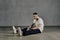 Athletic tattooed guy with beard. Dressed in white t-shirt, sneakers, black sports trousers. Sitting on floor, gray