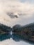 Atmospheric alpine landscape with trees near mountain lake and high snow-covered mountain in low clouds. Verical view