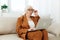 an attentive elderly woman in a brown suit is sitting at a laptop working remotely on the couch and peering at the text