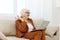 attentive elderly woman in a classic suit in a bright room on the sofa and working on a laptop remotely