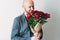 Attractive adult bald bearded man with funny face holding beautiful bouquet of dark peonies in craft paper on the grey background