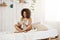 Attractive african girl in sleepwear sitting on bed smiling looking at tablet at home.