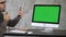 Attractive beard man checks his teeth on his phones selfie camera near computer monitor. Green Screen Mock-up Display.