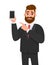 Attractive bearded businessman holding/showing brand new smartphone/mobile/cell phone in hand and pointing index finger towards.