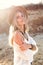 Attractive blonde girl with long hair is posing to the camera on beach. She wears white shirt with naked shoulsed, hat