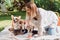 Attractive blonde girl sitting on white blanket in garden with cute welsh corgi puppies and dog.