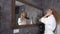 Attractive blonde woman in white bathrobe with long curly hair drying hair in front of mirror in bathroom at home