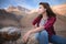 Attractive brooding young caucasian woman in gray T-shirt and jeans sits on a cliff against the mountains on a sunny day