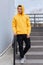 Attractive brunette guy standing on stairs in open air and looking dreamy away, young man dresses yellow hoodie, black pants and