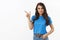 Attractive, coquettish female brunette in blue t-shirt inviting join company, pointing finger left to show advertisement