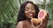 Attractive curly-haired african girl holding half of grapefruit close to her face looking at camera. Portrait of healthy