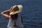 Attractive female person in white dress holding glamour hat outdoors on river shore, back view, deep blue water on background. cop