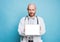 Attractive friendly bald bearded smiling doctor holds tablet with white screen looking at camera isolated on blue background