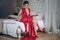 Attractive girl in evening red dress and red shoes in bedroom. the girl is sitting on the bed and straightening the dress. in a ch