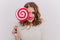 Attractive girl with gray eyes and bright lips is smiling, covering part of her face with lollipop on isolated