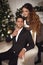 Attractive girl hugging handsome man on armchair. Romantic couple portrait. Cheerful Happy newlywed posing by xmas tree in Christ