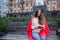 An attractive girl with long brown hair sits on a bench and writes her thoughts on the city background in a red notebook. She wear