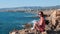 Attractive girl in red dress and sunglass sitting on edge of cliff and looking at sea. Happy girl sitting on rock and smiling. You