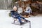 attractive girl rides sleigh in backyard. Snow-covered New Year house with fir trees and hearth with chairs