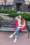 An attractive girl sits on a bench and writes her thoughts on the urban background in a red notebook. She wears a white sweater, b