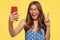 Attractive glad European woman with long hair, makes peace sign, holds mobile phone, poses for selfie, dressed in polka
