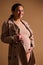 Attractive gravid woman, expectant mother in beige trench coat and underwear, smiling and gently stroking her bare belly