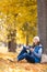 Attractive happy woman in warm wear sits under a tree with a mobile in hand and chill out in a autumn park outdoor