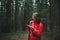 Attractive hiker girl walking along forest path in mountains and using smartphone, looking away. Hiker woman in red raincoat