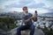 Attractive hipster with a beard is sitting on a rock high over Athens City with Acropolis view and makes a selfie