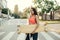 Attractive Hispanic girl walking with a longboard in her hands on the streets of an evening city, looking in camera, wearing