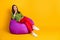 Attractive lady sit comfortable fluffy violet bean bag minded wear green sweater red pants sneakers isolated yellow