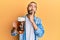 Attractive man with long hair and beard drinking a pint of beer serious face thinking about question with hand on chin, thoughtful