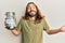 Attractive man with long hair and beard holding charity jar with money celebrating achievement with happy smile and winner