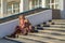 Attractive middle-aged woman wearing stylish coat and shoes sitting with bag on stairs step of office building in early spring at