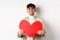 Attractive modern man smiling and looking at camera hopeful, holding big red Valentines heart, waiting for soulmate on