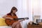 Attractive musician woman sitting in the living room playing acoustic guitar and singing songs. Teenager holding acoustic