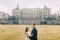 Attractive newlywed couple embracing at green sunny lawn near beautiful ruined baroque palace. Loving groom holding