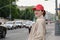 Attractive red-haired young woman in a red baseball cap and beige raincoat stands on the side of a city road