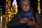 Attractive senior lady messaging with the cellphone. Outdoor in the night with a Christmas tree behind her. Yellow lights