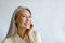 Attractive silver haired woman in beige shirt touches cheek on light grey background