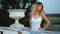 Attractive skinny girl in white and blue dress stands near white stone balustrade with flowerpot, looks aside with hands