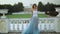 Attractive skinny girl in white and blue dress stands near white stone balustrade smiling, dancing and posing during
