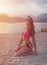 Attractive slim young brunette woman in bikini and sunglasses sitting on beach smiling admiring sunrise with mountains