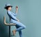 Attractive smiling short haired brunette woman in blue business smart casual suit, shoes, hat sitting on stool looking