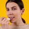 Attractive smiling young woman using hygienic lipstick on yellow background. lips care and protection. woman applying balm on lips