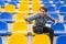 Attractive sporty young man model in blue shirt sitting on blue stadium seats after training staring at field