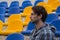 Attractive sporty young man model in blue shirt sitting on blue stadium seats after training staring at field