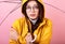 Attractive student in a yellow raincoat with a hood on her head with glasses under an umbrella, snuggles her hands because of the