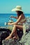 Attractive tanned girl in hat sitting on the stoned beach and making a drink, holding kettle from propane stove and cup.