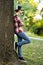 Attractive teenage boy in checkered shirt, blue jeans, sunglasses and baseball cap lean on a tree waiting for someone or